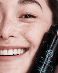 woman holding a black bottle labeled 'Bella Aura cleanser' near their face.